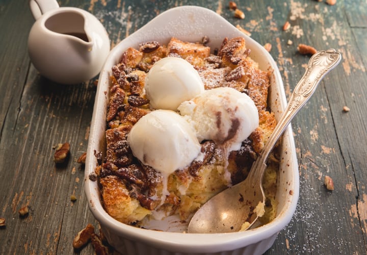 bread pudding with a piece missing, a spoon in the pan with 3 scoops of vanilla ice cream on top