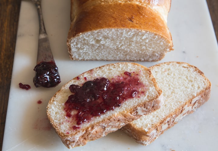 jam on a slice of milk bread on a white board with the loaf and a knife