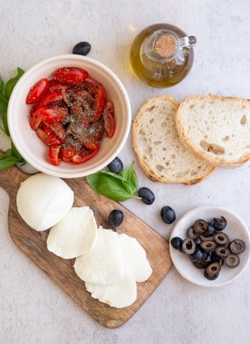 ingredients for bruschetta, bread, tomatoes in a bowl, sliced mozzarella on a board and olives