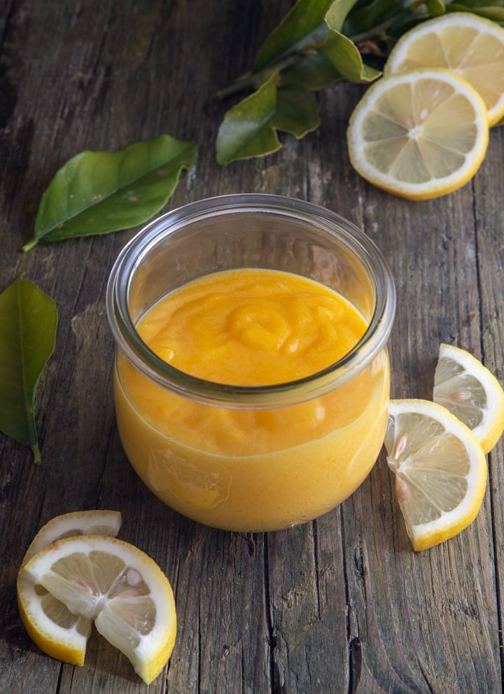 lemon curd in a glass jar with sliced lemons and leaves on a board.