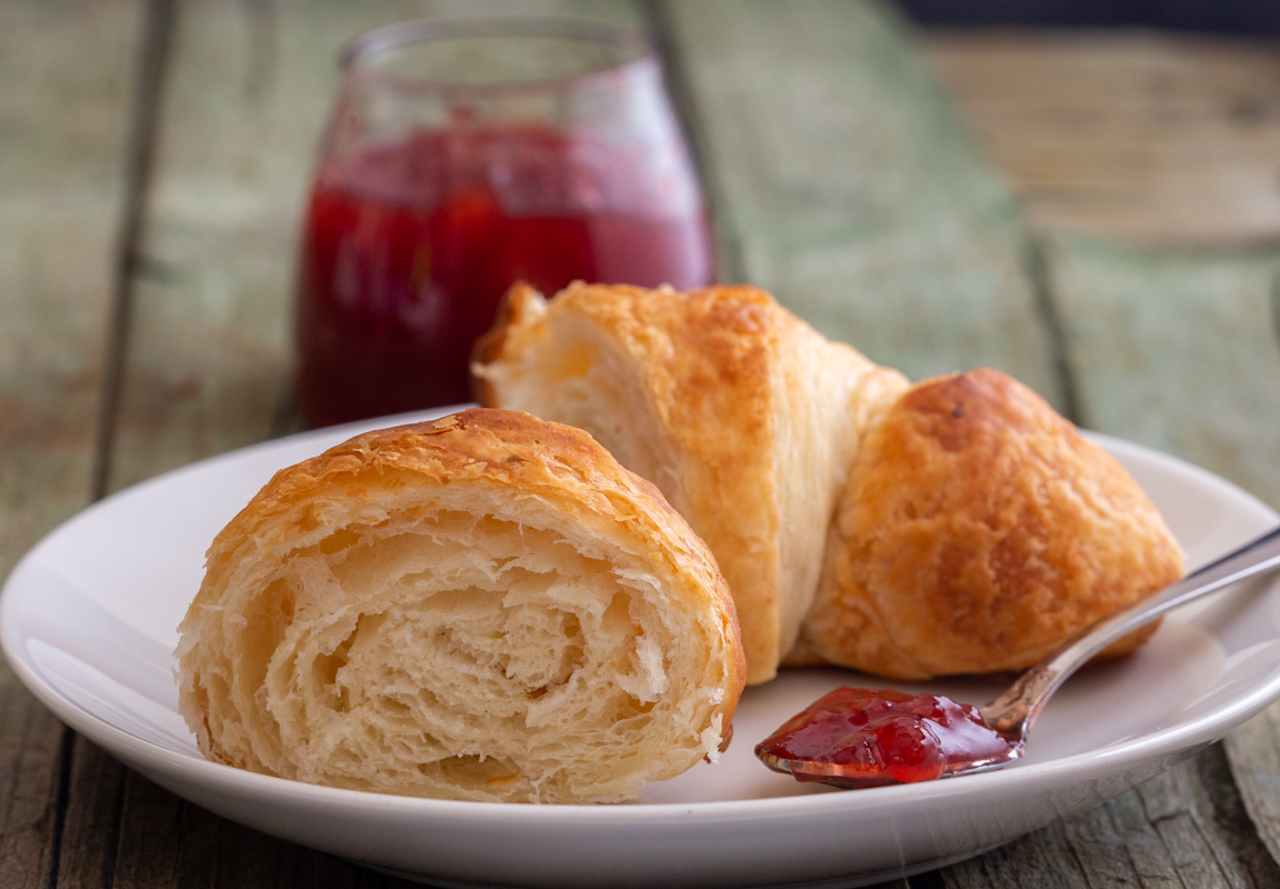 A croissant sliced in a half on a white plate with jam on a spoon.