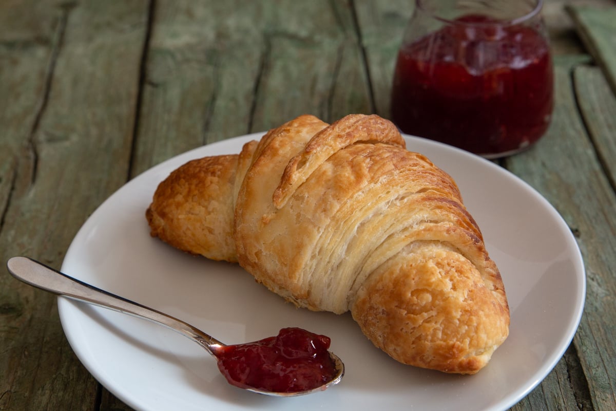 Croissant on a white plate with jam on a spoon.