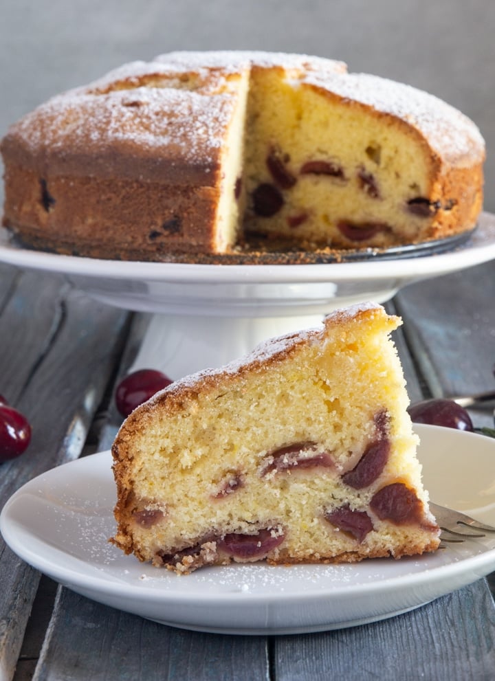 Cherry cake on a white cake stand and a slice on a plate.