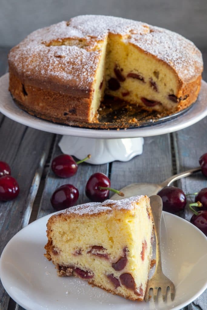 Cherry cake on a white cake stand and a slice on a plate.
