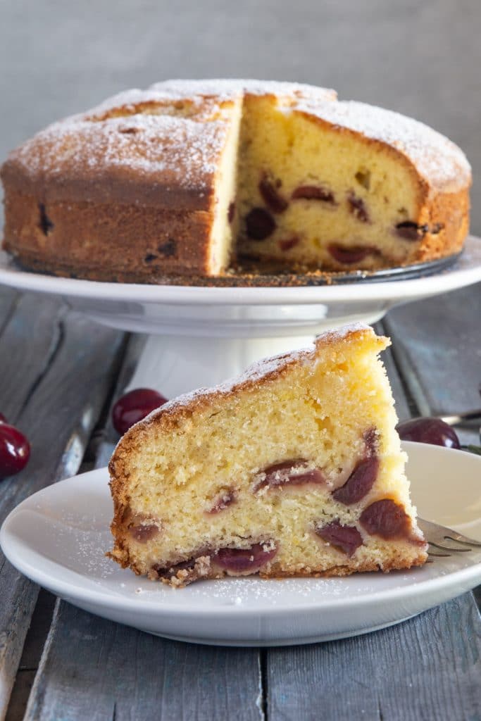 Cherry cake on a white cake stand and a slice on a white plate.