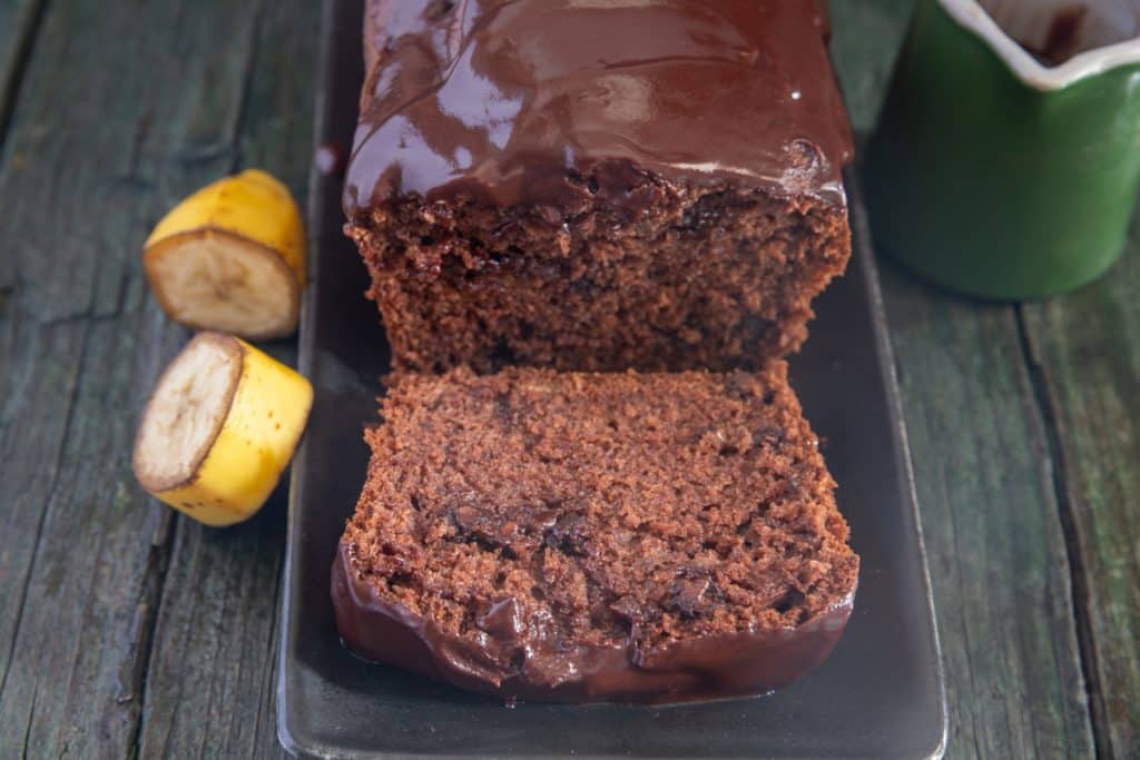 Chocolate banana bread cut with a slice in front on a black plate, two pieces of banana on the left side and a green mug on the right side.