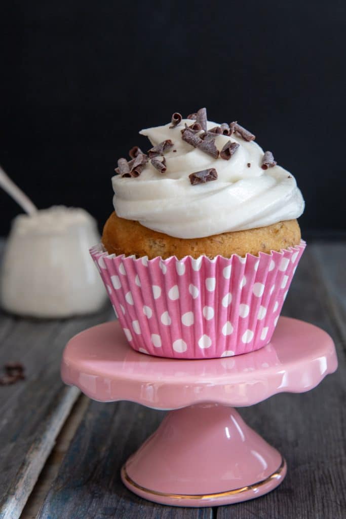 A cupcake on top of a small pink cake stand with some frosting in a jar in the back.