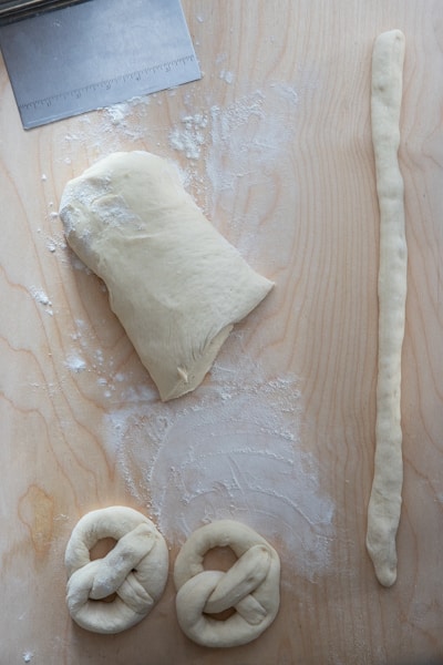 Two pretzels made on a wooden board with the rest of the dough cut and a rope ready to be formed into a pretzel.