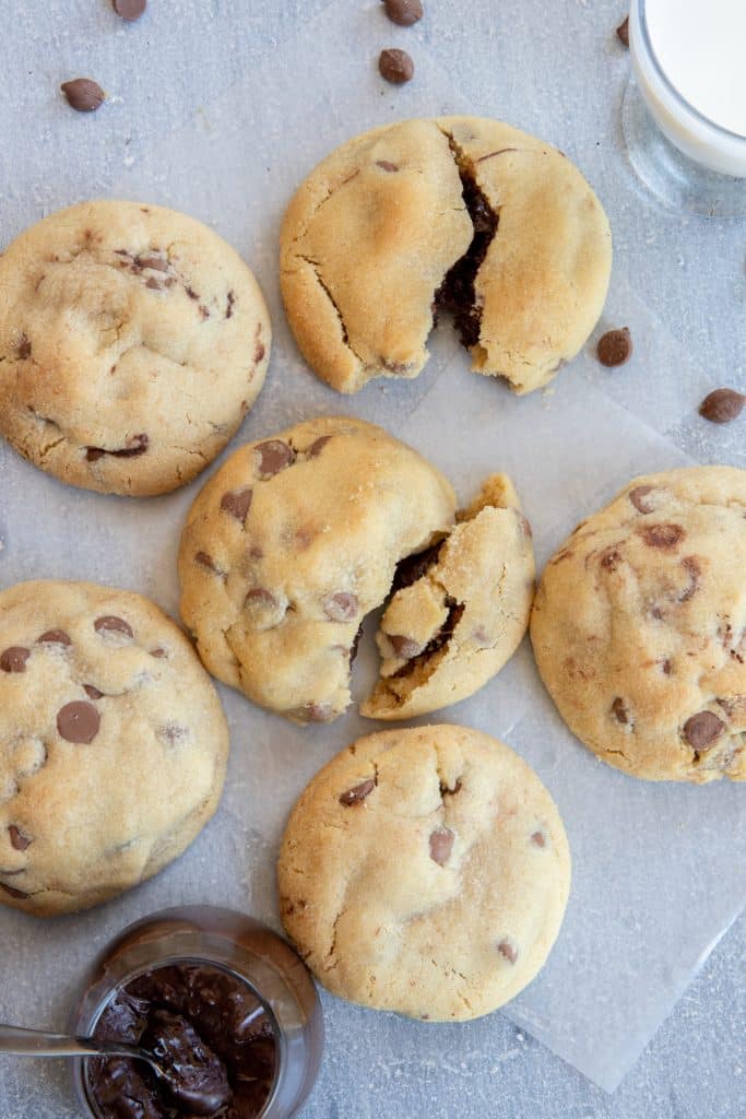 six cookies lying on a piece of parchment paper with some chocolate chips scattered around and a small jar of chocolate spread on the side.
