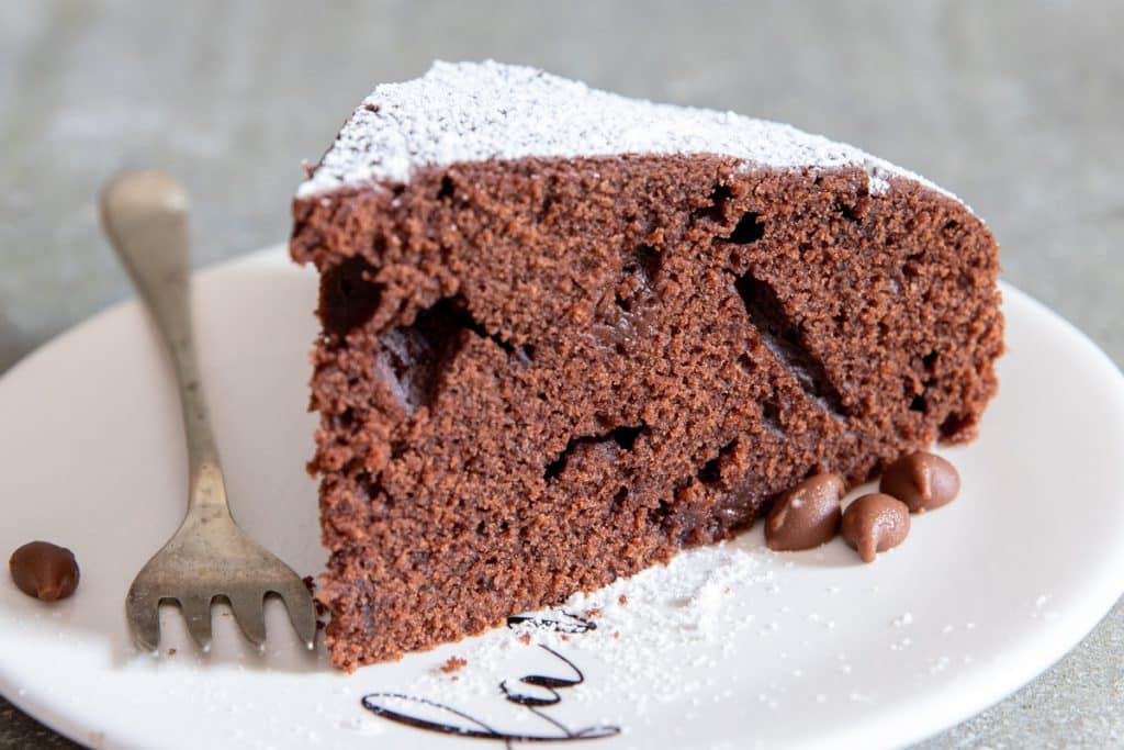 A slice of cake on a white plate with a fork on the side and chocolate chips scattered around.