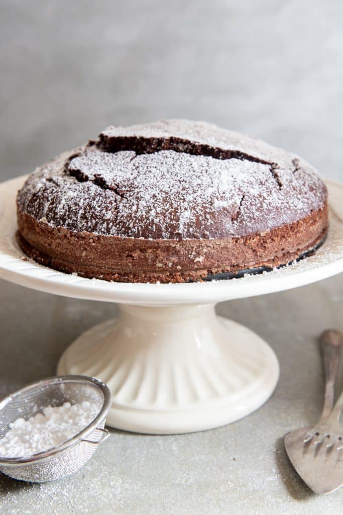 cake on a white cake stand with powder sugar in a sifter and a fork on the side.