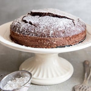cake on a white cake stand with powder sugar in a sifter in front.