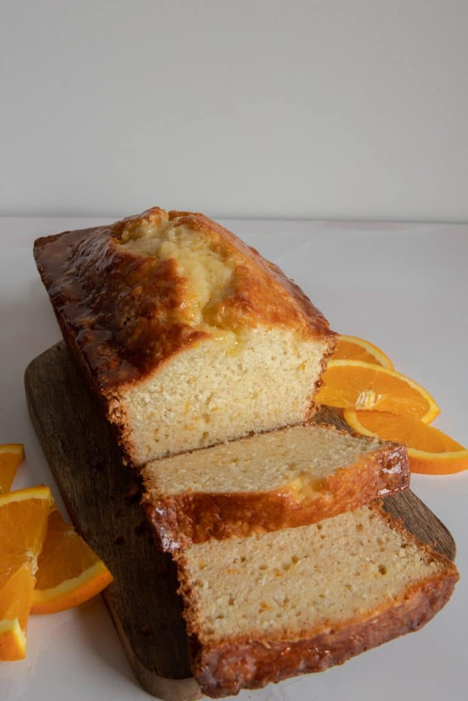 orange bread on a wooden board with two slices in front and orange slices on the sides.