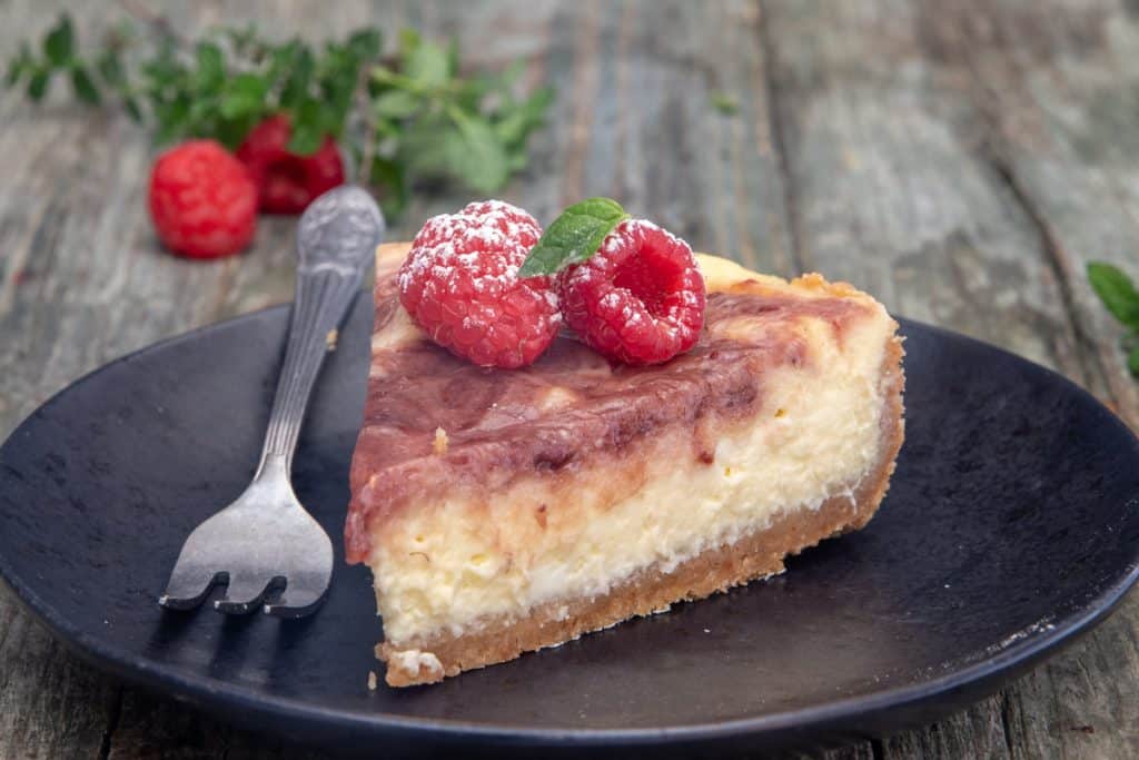 A slice of cheesecake on a black plate with raspberries on top and a fork on the left side.