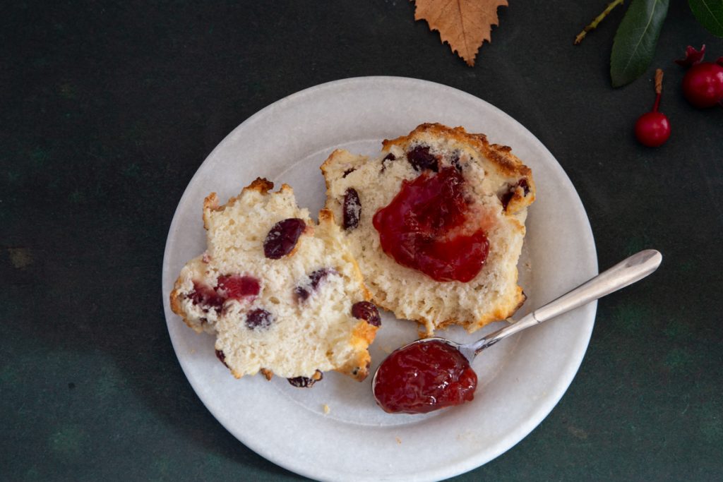 A biscuit cut with jam on a white plate with some jam on a silver spoon.