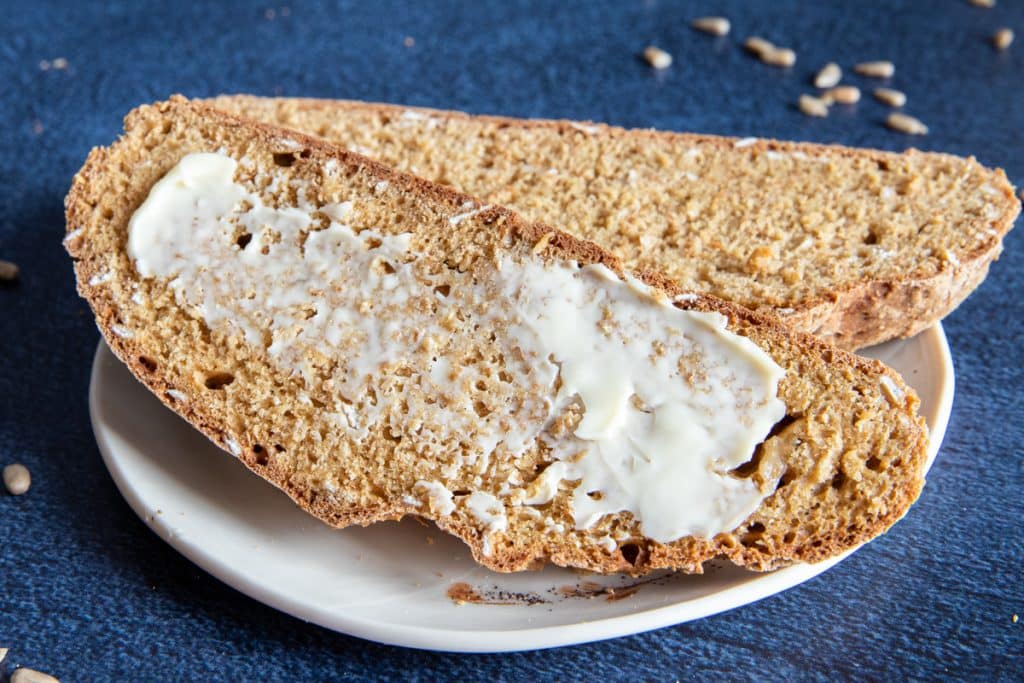 Two slices of brown soda bread on a white plate with one slice buttered.