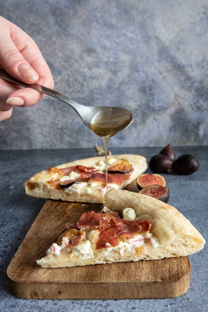 Two slices of pizza on a wooden board with a hand drizzling honey with a spoon on one of the slices.