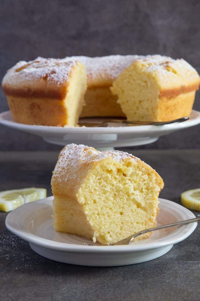A slice of lemon cake on a white plate with the rest of the cake on a cake stand in the back.