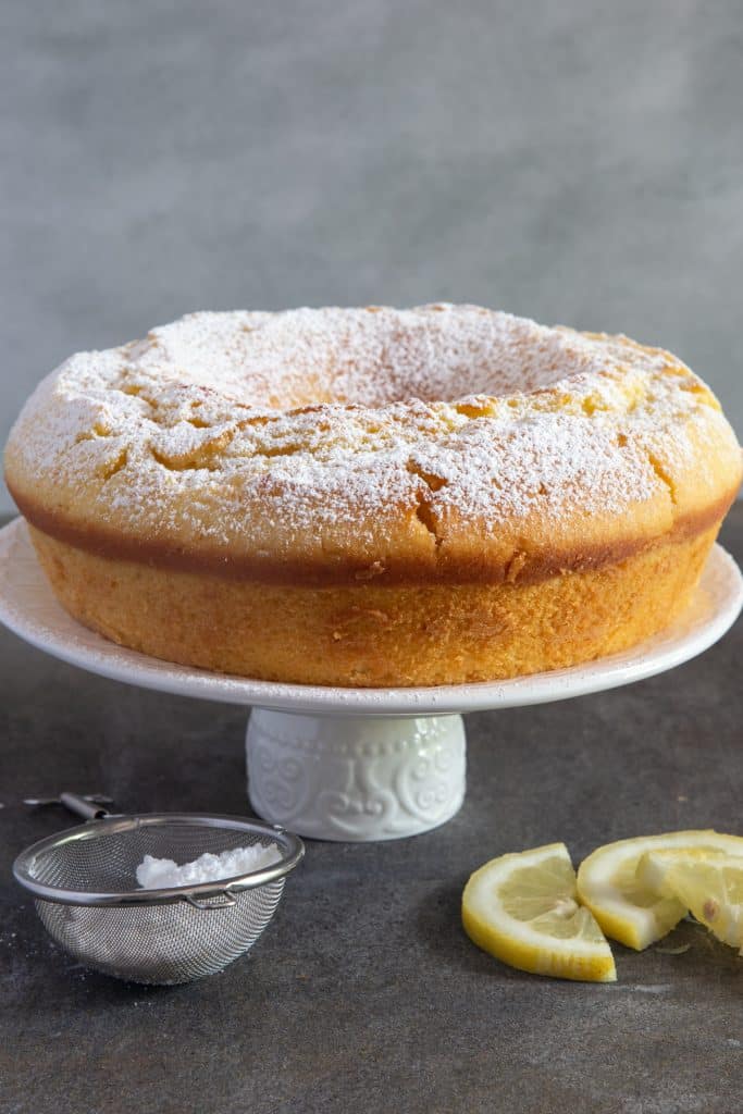 Lemon cake on a cake stand with two slices of lemon on the right side and powdered sugar in a sifter on the left side.