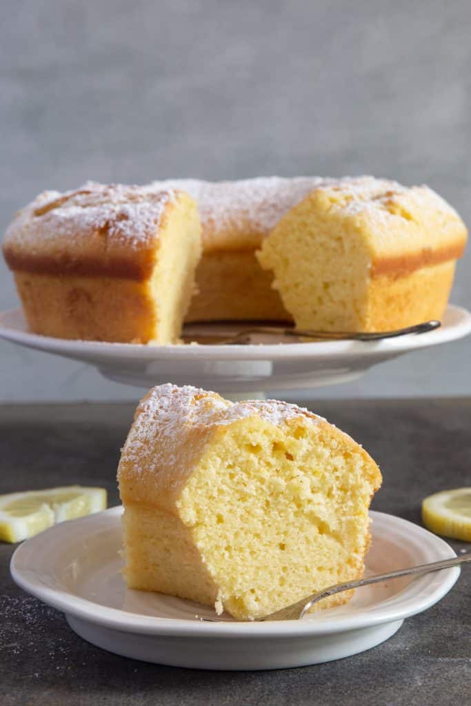Slice of lemon cake on a white plate with cake in the back on a stand.