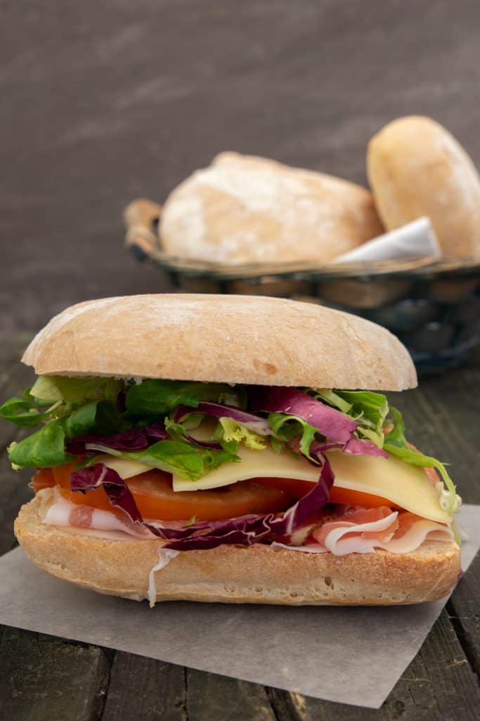 A sandwich on a piece of parchment paper with a basket of bread buns in the back.