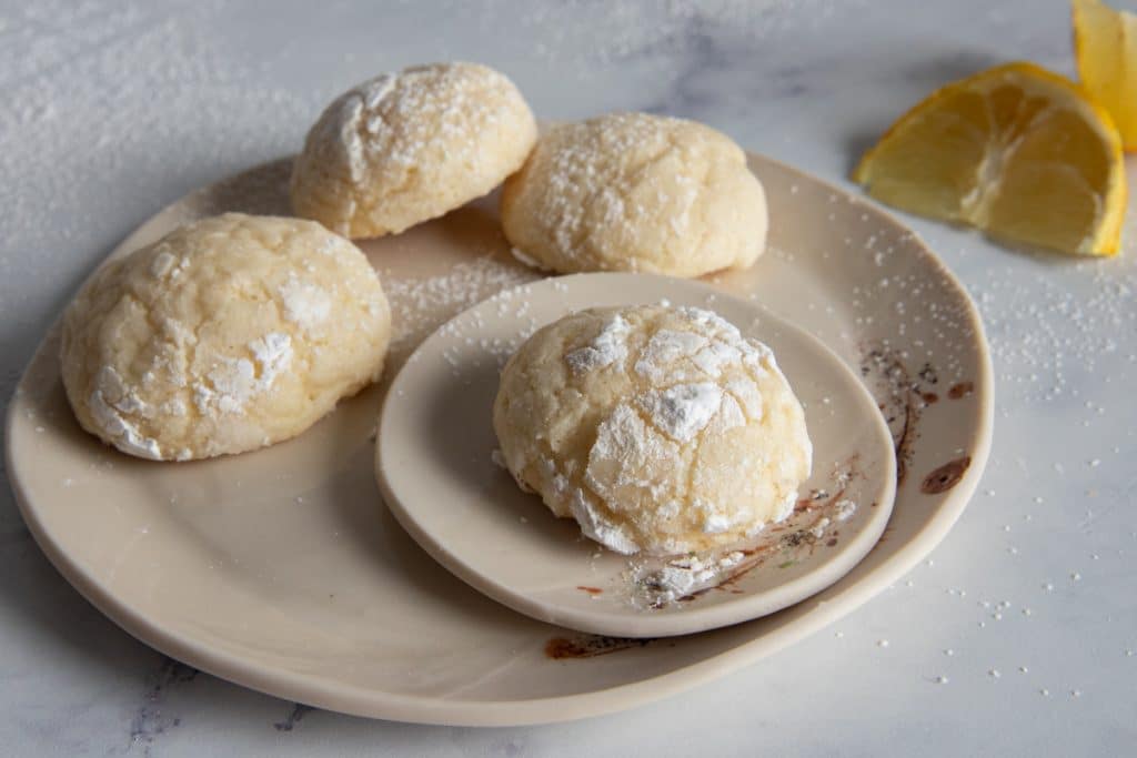 Four lemon cookies on a white plate with two lemon slices in the back.