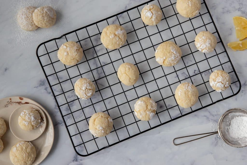 Cookies on a black wire rack with a white plate with three cookies on top on the left side and powdered sugar in a sifter on the bottom right side.