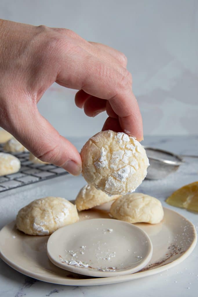 One lemon cookie held by a hand with three more cookies on a white plate.