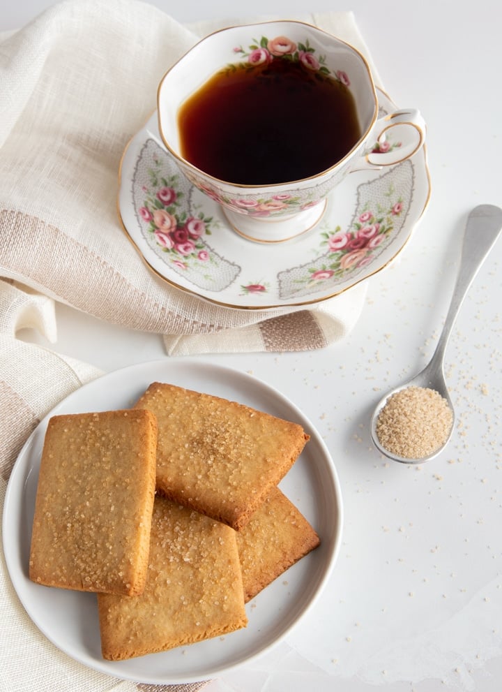 Four tea cookies on a white plate with a spoon filled with brown sugar on the right side and a cup of tea above the cookies.