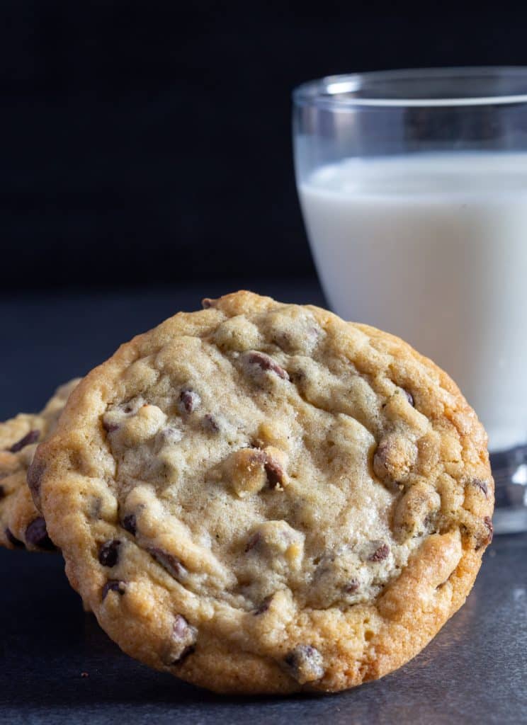 One chocolate chip cookie with a glass of milk in the back.