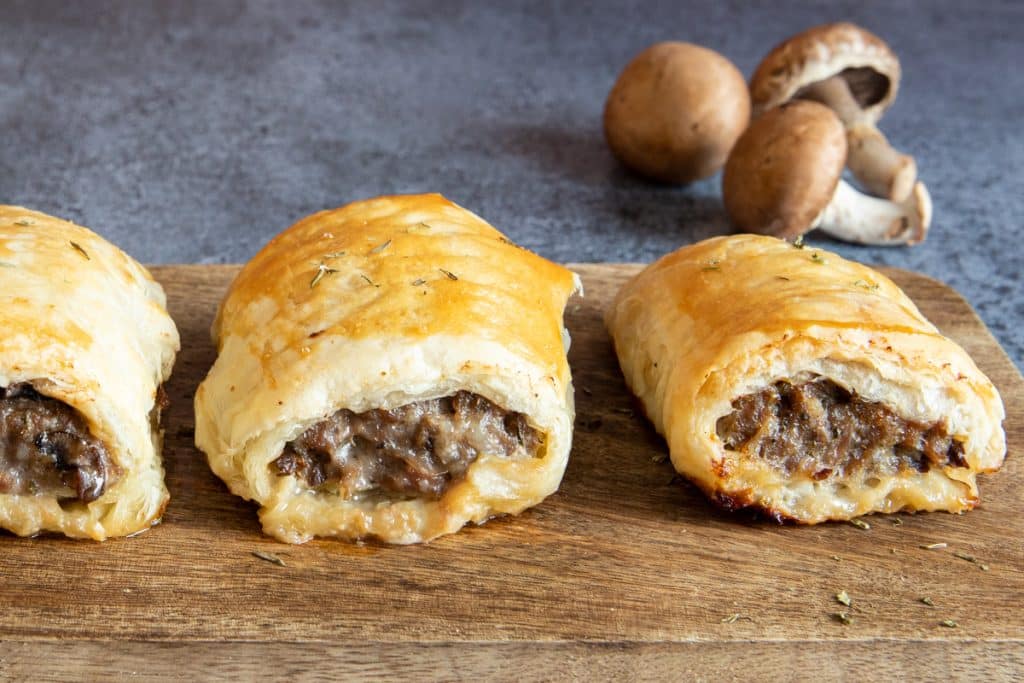 Three sausage rolls on a wooden board with three brown mushrooms in the back.