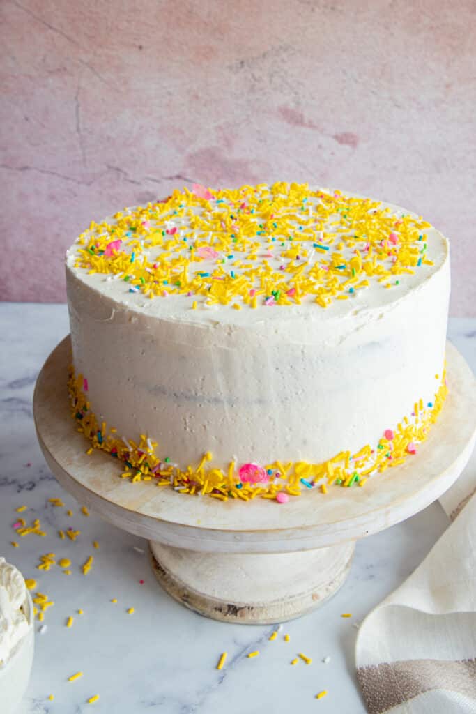 Fudge cake on a white cake stand with yellow sprinkles around it and a white cloth on the right side.