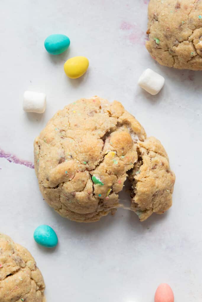 A broken chunky cookies with two other cookies around it and marshmallows and mini eggs scattered around.