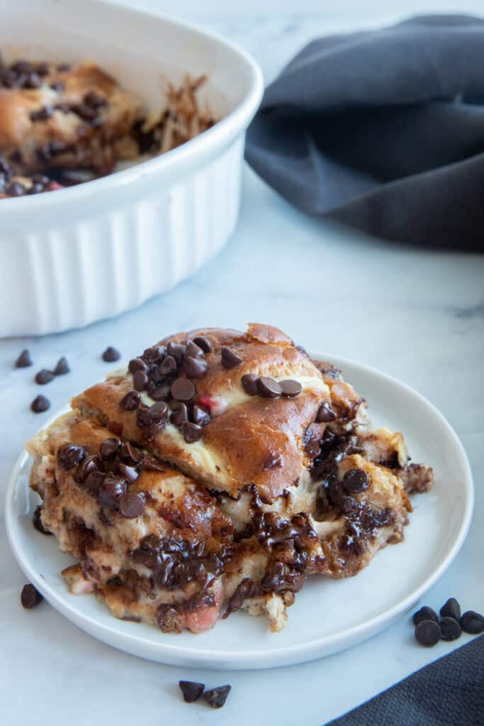 A piece of bread pudding on a white plate with the rest in a baking sheet in the back and chocolate chips scattered around.