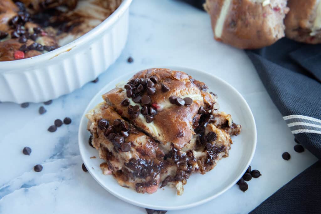 One piece of bread pudding on a white plate with two hot cross buns on the side and a black cloth under the buns.