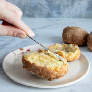 A scone cut in half an buttered by a hand holding a butter knife.