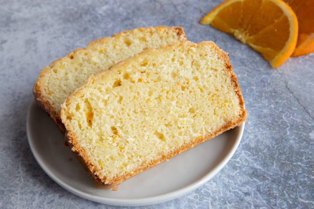 Two slices of madeira cake on a white plate with two slices of orange on the side.
