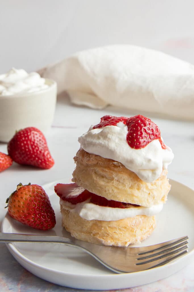 Two biscuits topped with whipping cream and strawberries on a white plate with three more strawberries on the left side of the plate.