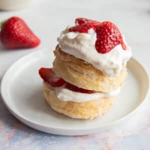 Two biscuit on top of each other topped with whipping cream and sliced strawberries on a white plate.