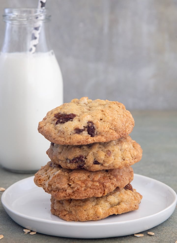 Four oatmeal raisin cookies stacked on a white plate and a glass bottle of milk behind them.