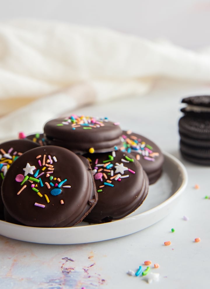 Five chocolate covered oreos on a white plate with three oreo cookies stacked on the right side and sprinkles scattered around.