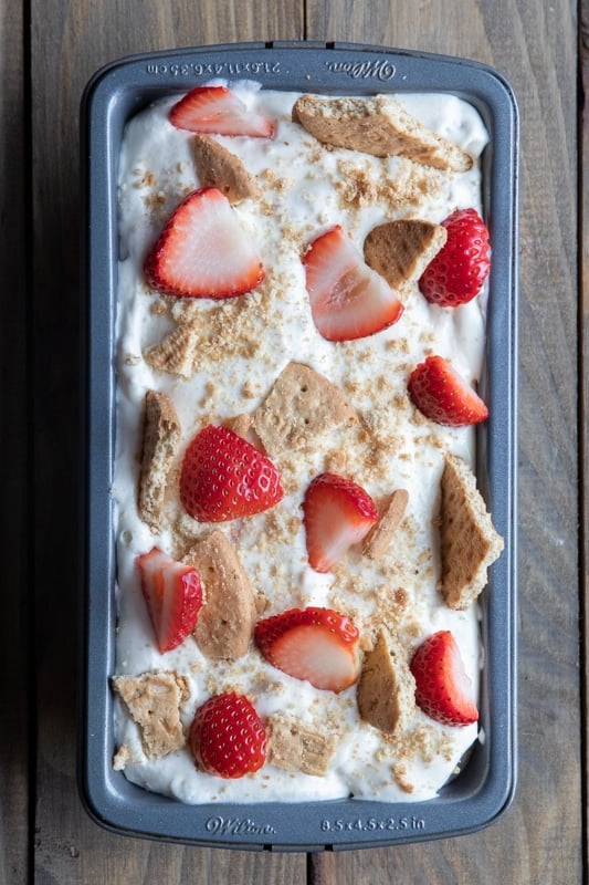 Ice cream in a loaf pan with sliced strawberried and broken cookies on top.