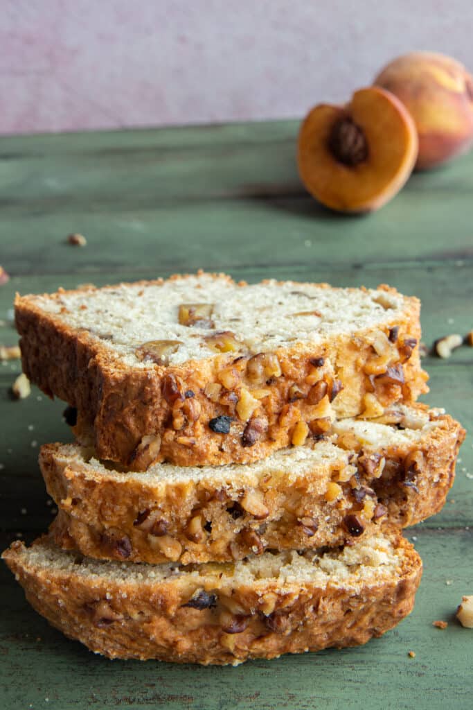 Three slices of peaches and walnuts bread one on top of the other with a peach cut in half in the back.