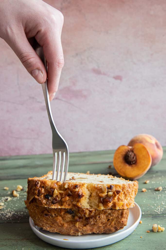 A hand holding a fork over a slice of peach bread on top of another slice no a white plate.