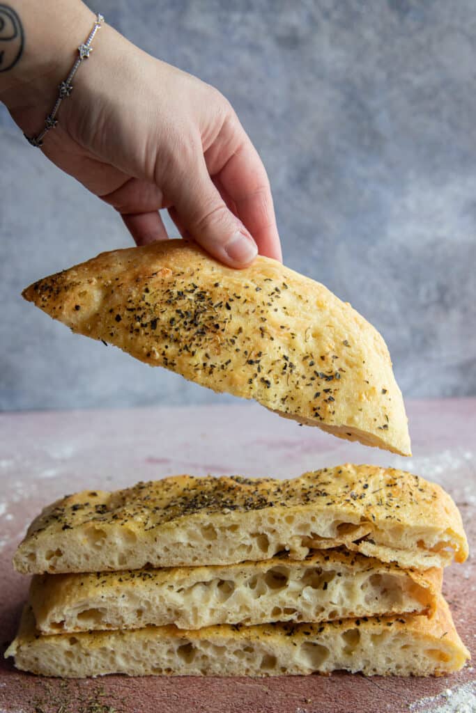 A hand holding a slice of focaccia and three other slices on top of each other.