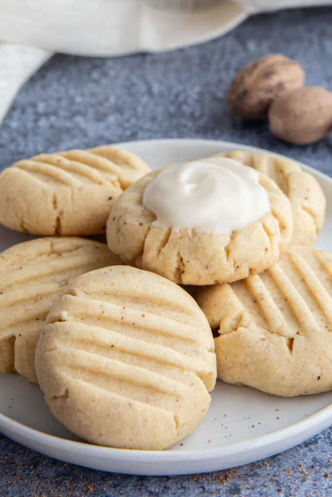 Five nutmeg cookies on a white plate with a white cloth on the left top side and two nutmegs on the top right side of the photo.