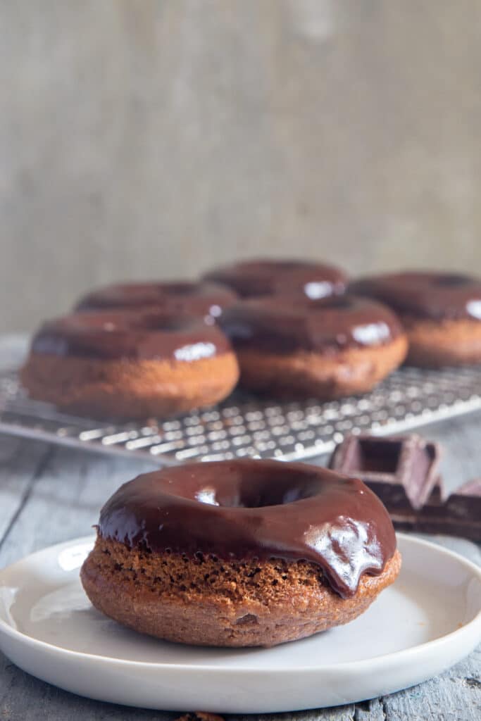 A chocolate donut on a white plate with five more donuts on a cooling rack behind it.