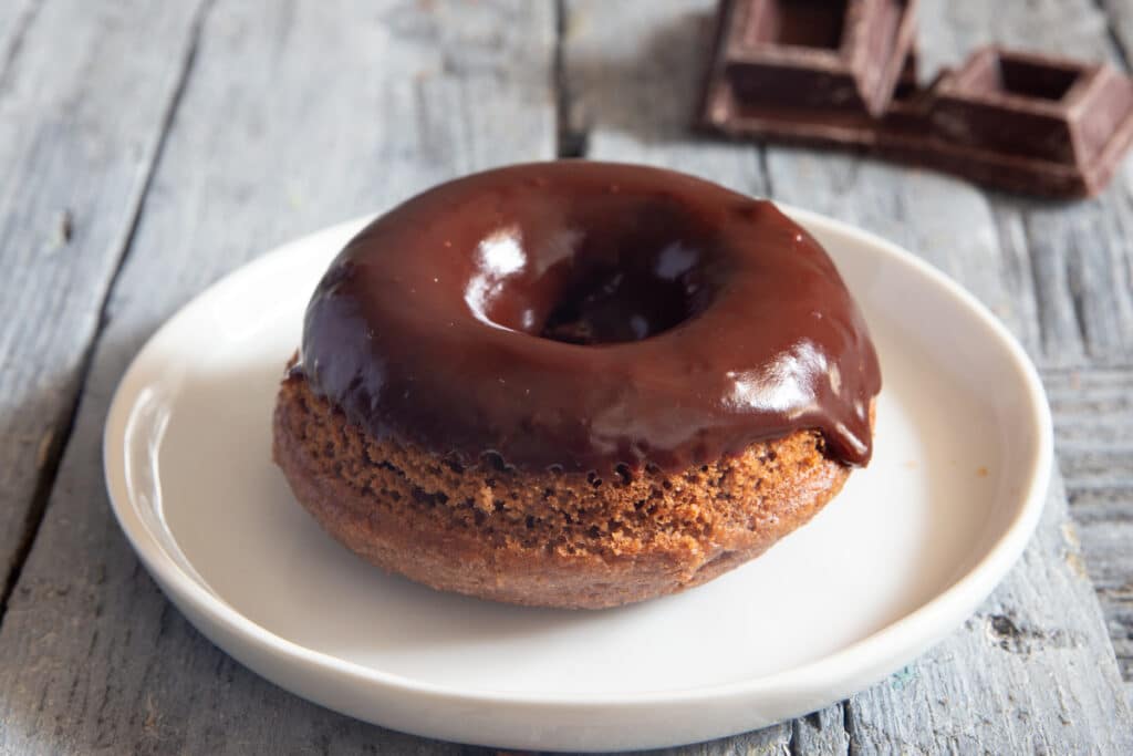 One chocolate glazed donut on a white plate with two chocolate squares behind the plate.