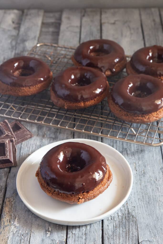 One chocolate glazed donut on a white plate with five more donuts on a cooling rack.