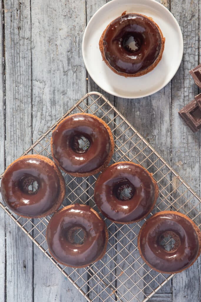 A donut on a white plate and five more chocolate donuts on a cooling rack.